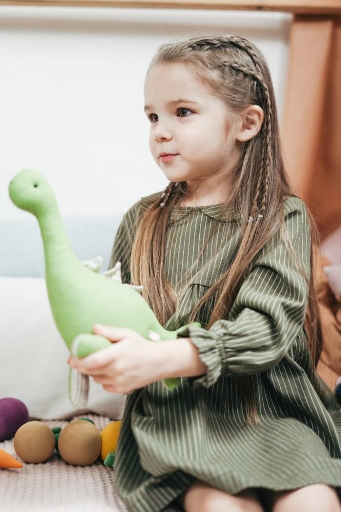 Adorable young girl in a green dress holding a charming stuffed dinosaur toy indoors.